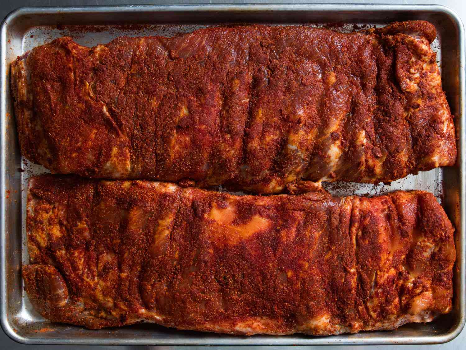 Two racks of pork ribs coated in dry rub on a rimmed baking sheet