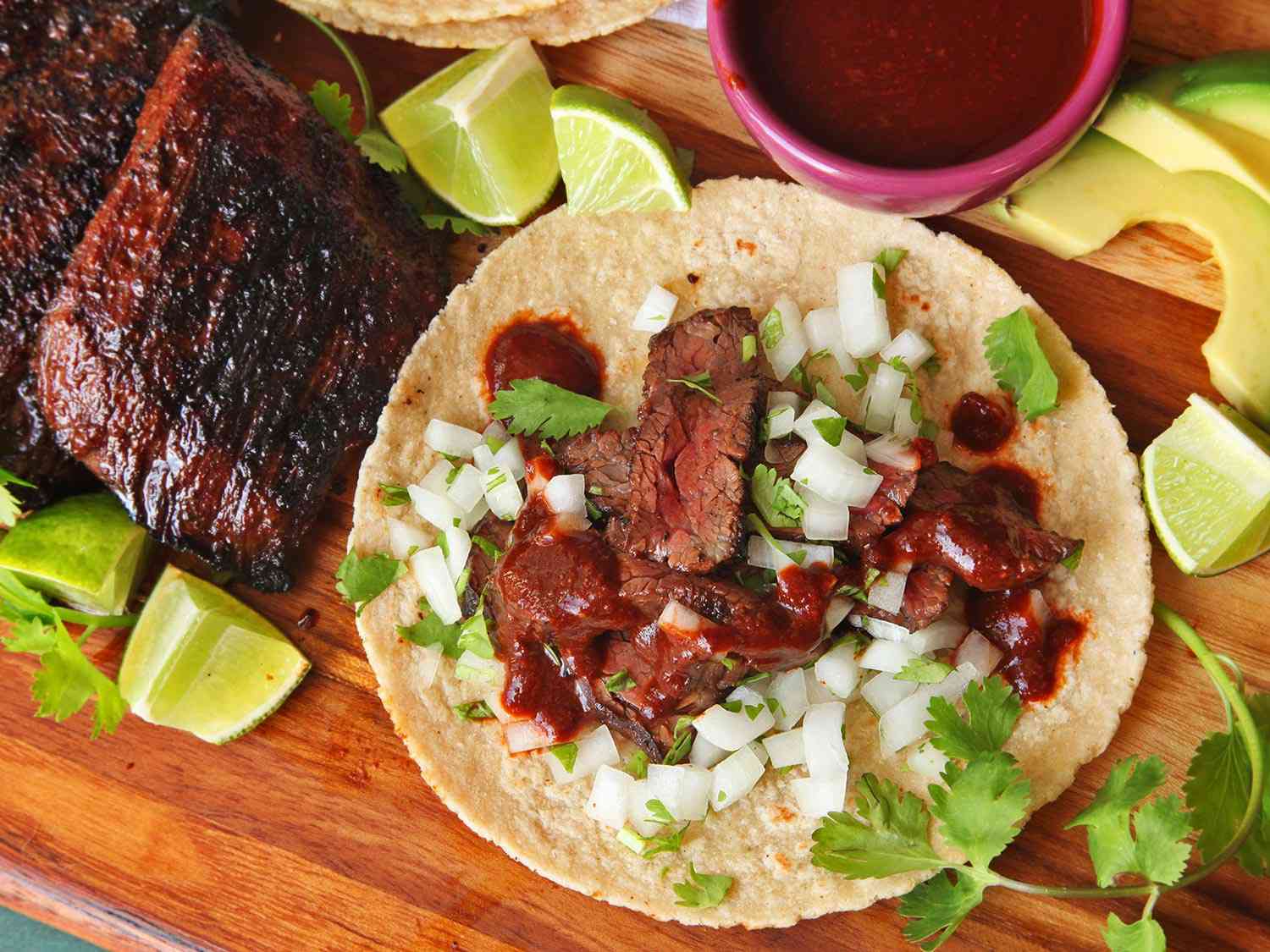 A carne asada taco, made with grilled skirt steak, on a wooden cutting board with additional steak, limes, avocados, cilantro, and salsa surrounding it.