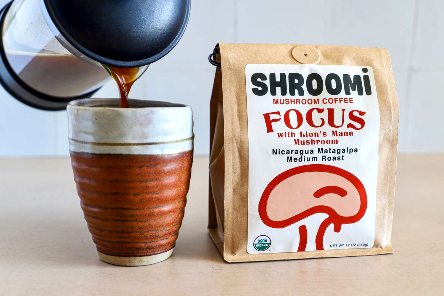 A cup being filled with coffee from a French press beside a bag of Shroomi Focus mushroom coffee
