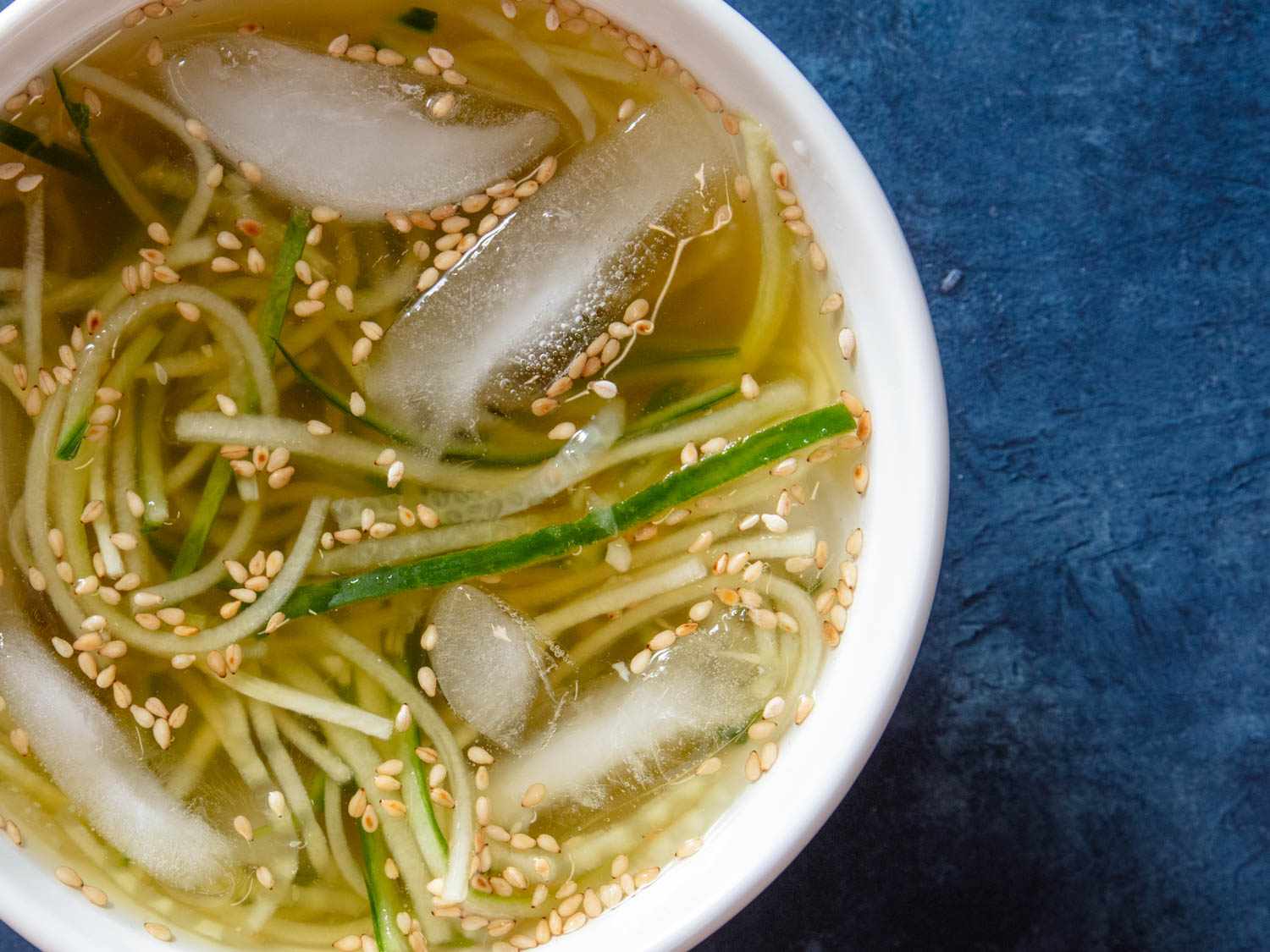 Overhead closeup of a bowl of oi naengguk. Ice cubes and sesame seeds float on the surface of the broth.