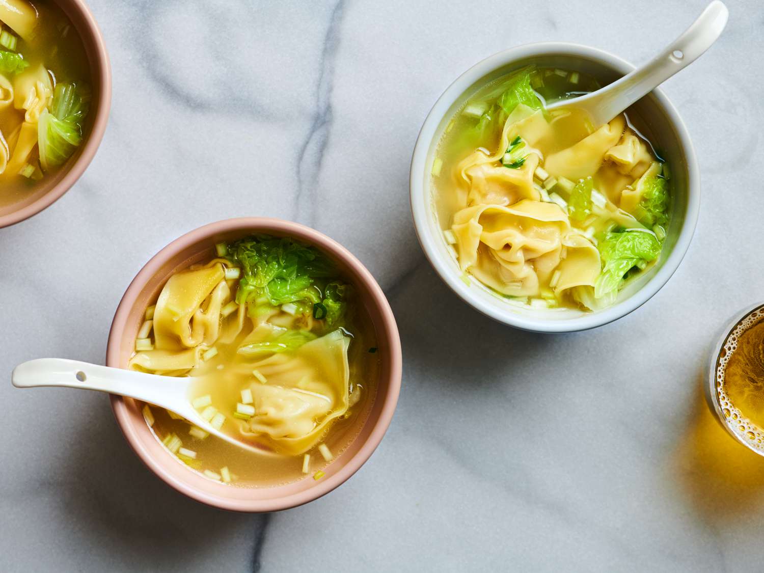 Two bowls of wonton soup on a marble surface. Each bowl has a white ceramic soup spoon in it. There is another bowl on the top left corner of the image, and a glass of liquid on the bottom right periphery.
