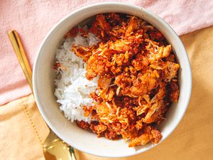 A bowl of white rice topped with a spiced chicken dish, served alongside a gold utensil on a cloth background