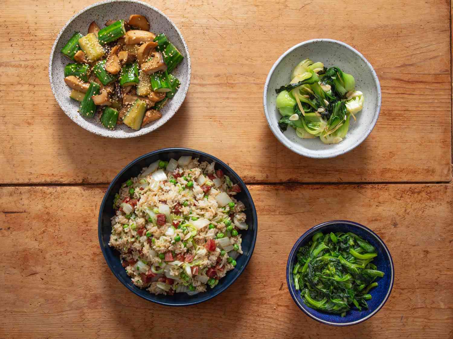 Overhead of four dishes made using the torch hei technique: stir-fried cucumbers with mushrooms, fried rice with Chinese sausage and cabbage, stir-fried bok choi, and yu choi.