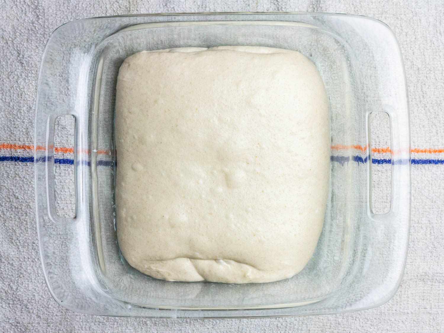 The raw dough for sourdough bread sits in a square glass baking dish; it looks soft and tender with some visible pockets of air under the surface.