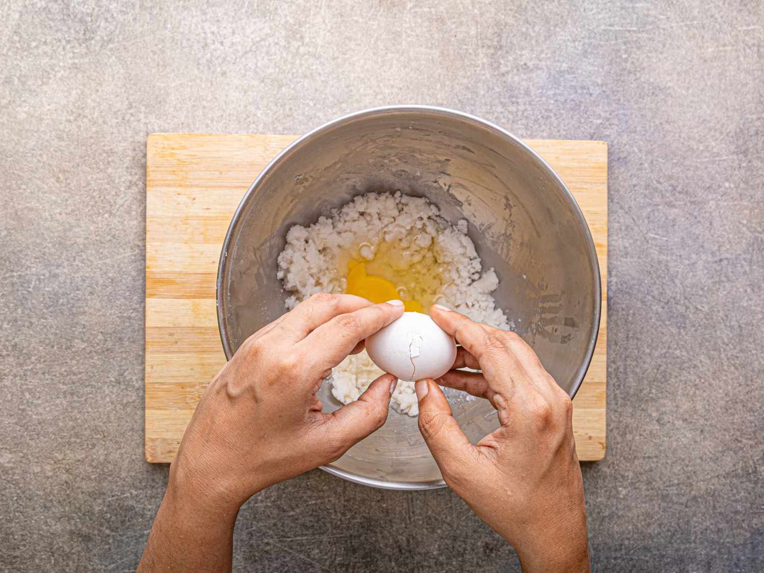 A persons hands cracking an egg into a mixing bowl containing shredded coconut on a wooden cutting board surface