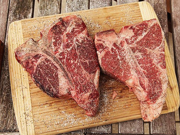 Two salted porterhouse steaks being sprinkled with ground black pepper.