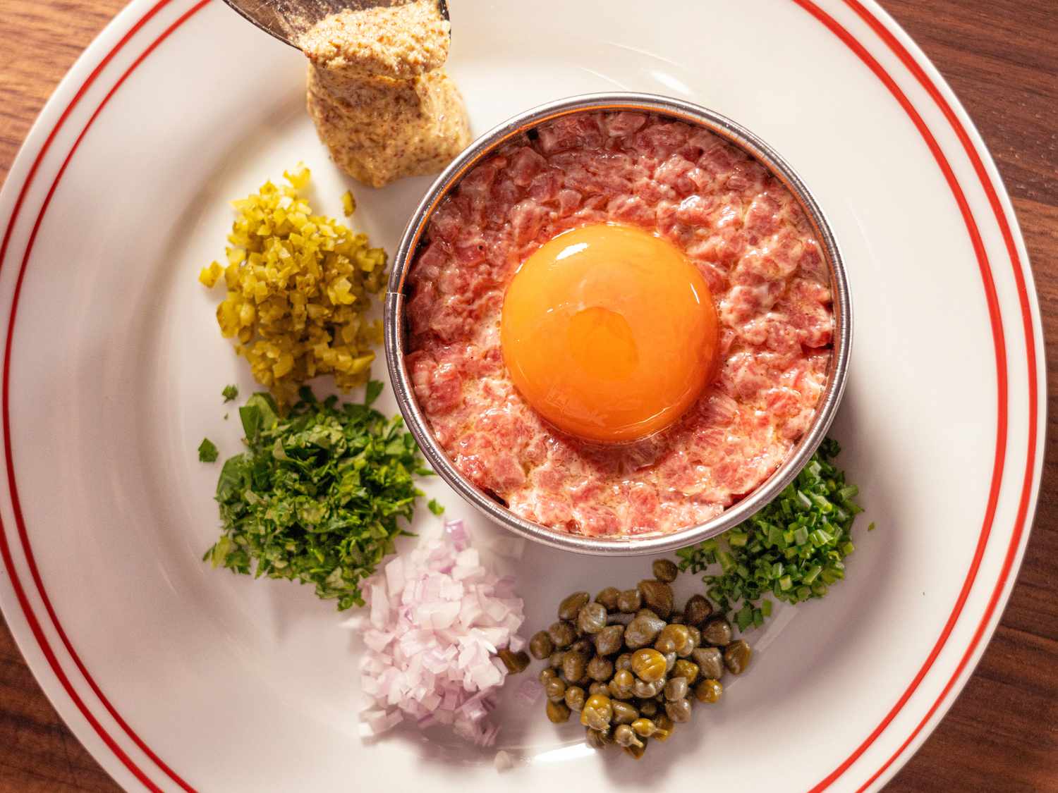 beef tartar on a dish with a raw egg dropped into form, and capers, onions, mustard and herbs stacked around it on a white plate 