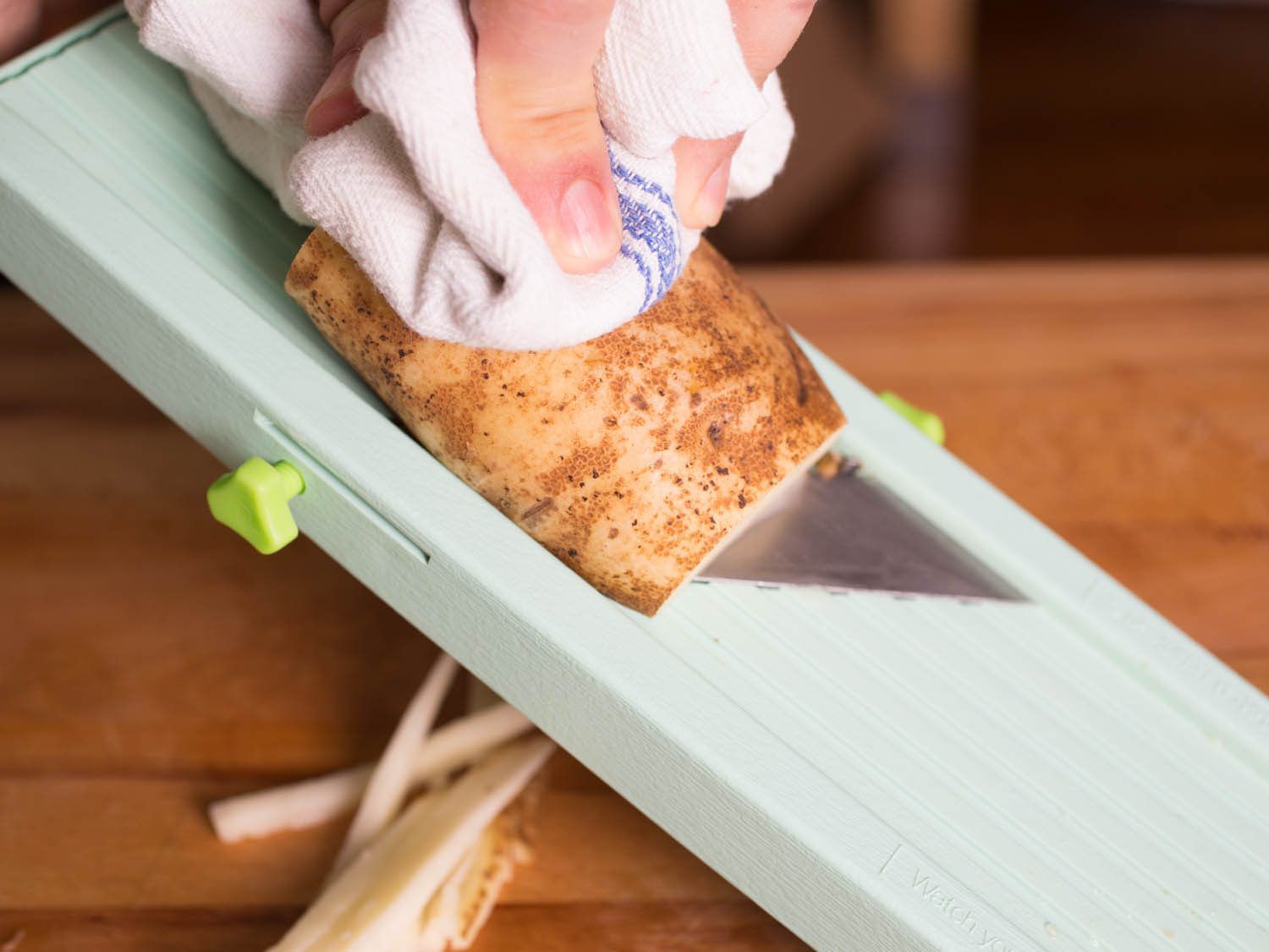 A potato slicing on a mandoline. A hand is holding the potato with a kitchen towel in between. 