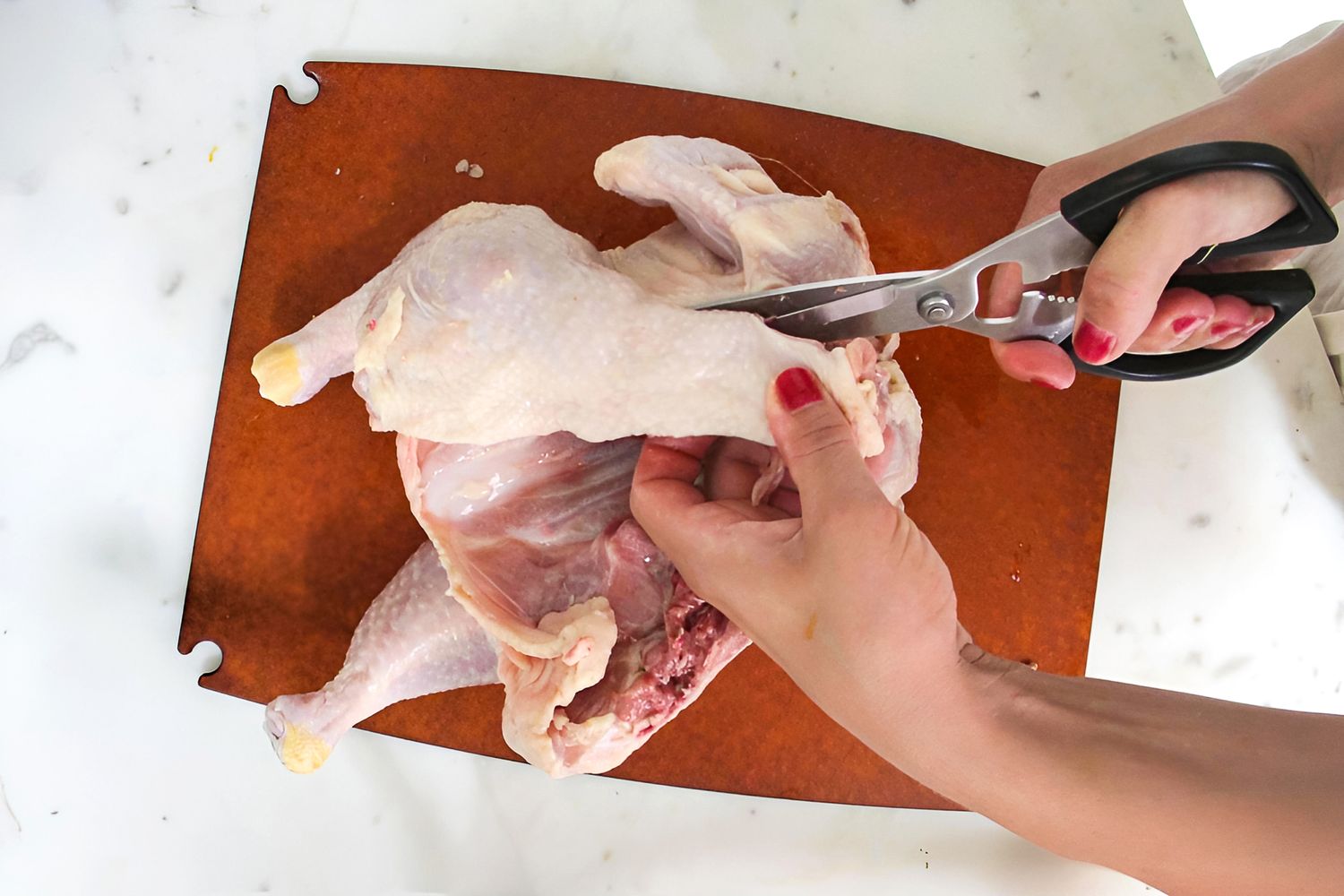 Hands using kitchen shears to cut a whole raw chicken on a cutting board