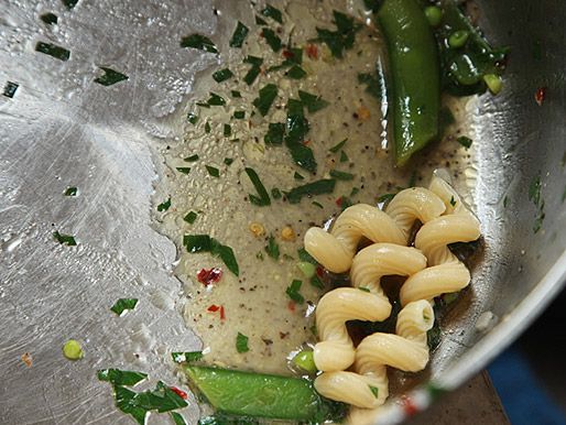 Closeup of pieces of pasta and snap peas in the bottom of a saucepan. A "broken" sauce of olive oil and pasta cooking water is pooled around them.