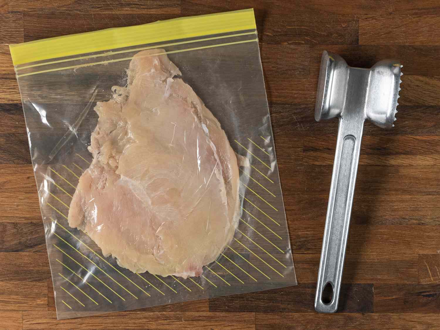 Chicken breast inside of a sealed zipper-lock bag, pounded to an even one-quarter inch thickness, with a metal meat pounder next to the bag. The bag and hammer are both on a wooden background.