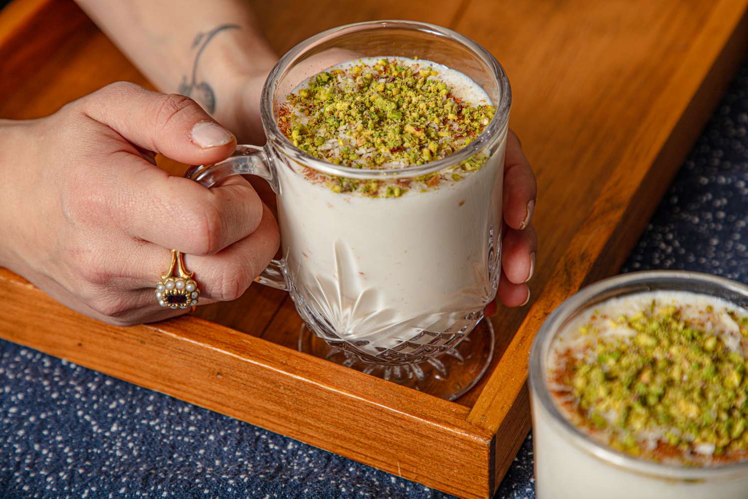 A hand holding a glass of sahlab topped with chopped pistachios on a wooden tray