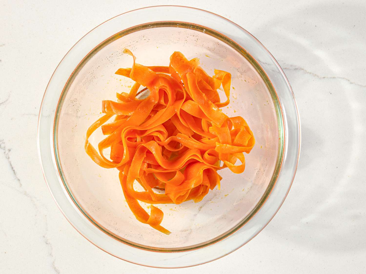 A glass bowl containing carrot ribbons on a light surface