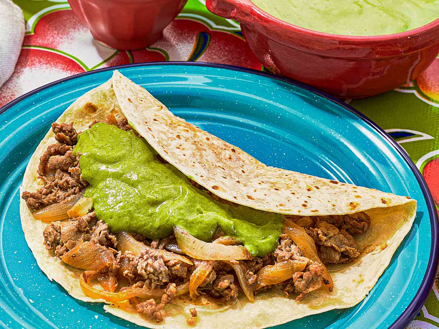 taco with mean topped with guacamole, with bowl of guacamole. Plate of tortillas, and 2 cocktails, and small dishes of onions and limes, on colorful floral printed tablecloth