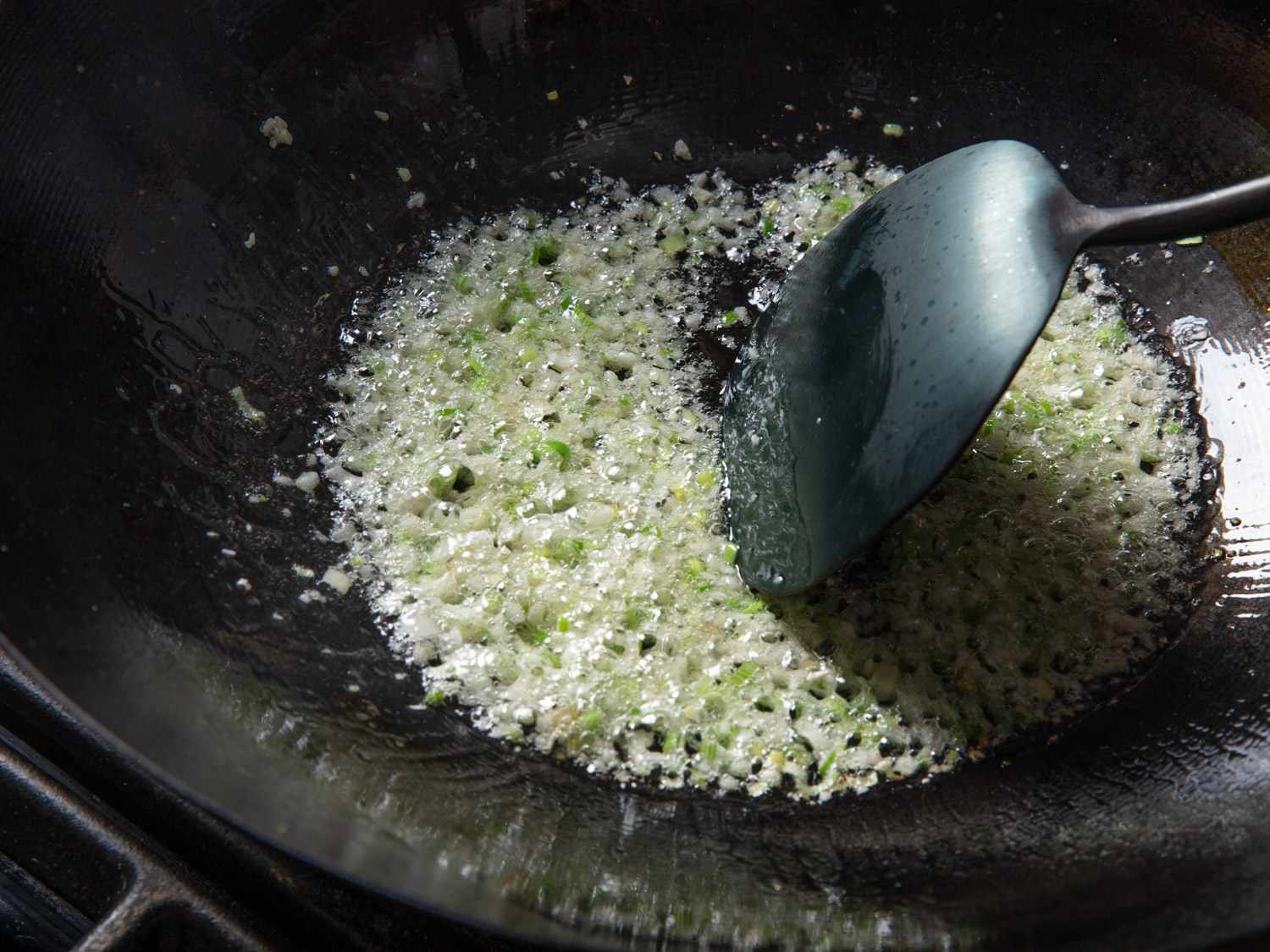 A metal spatula stirring minced garlic and scallion whites in a well-oiled wok. 