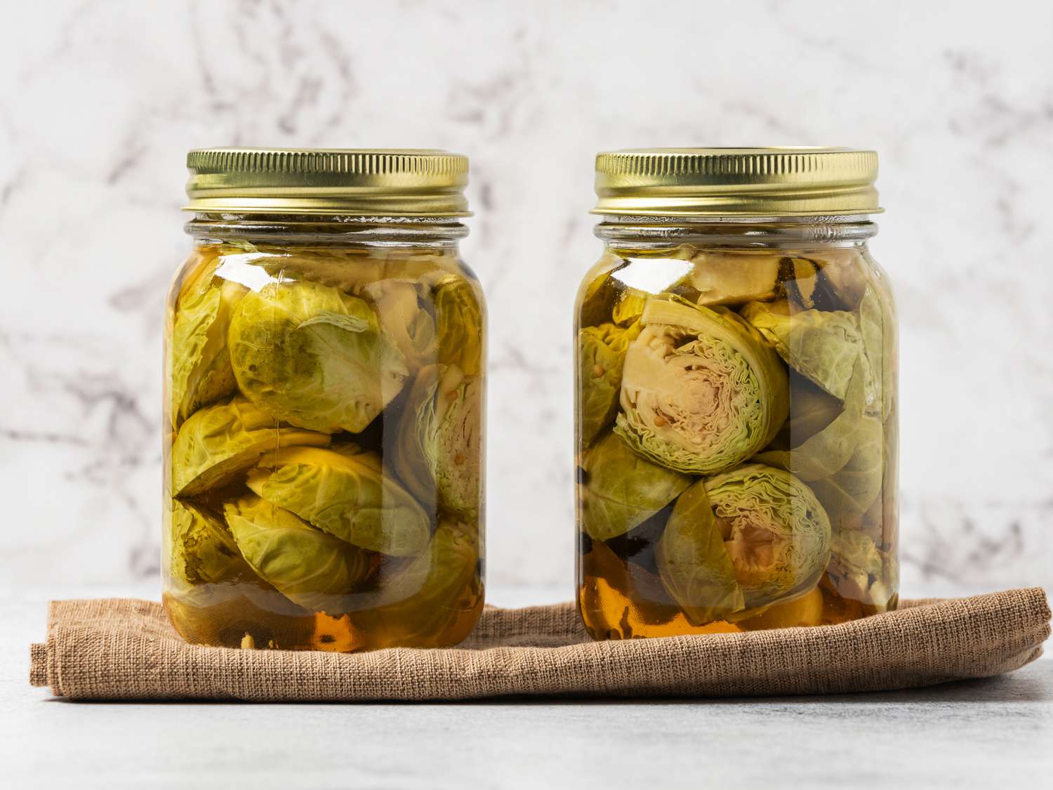 The two fully processed jars of Brussels sprouts placed on top of a folded brown cloth napkin. The jars are in front of a white marbled background.