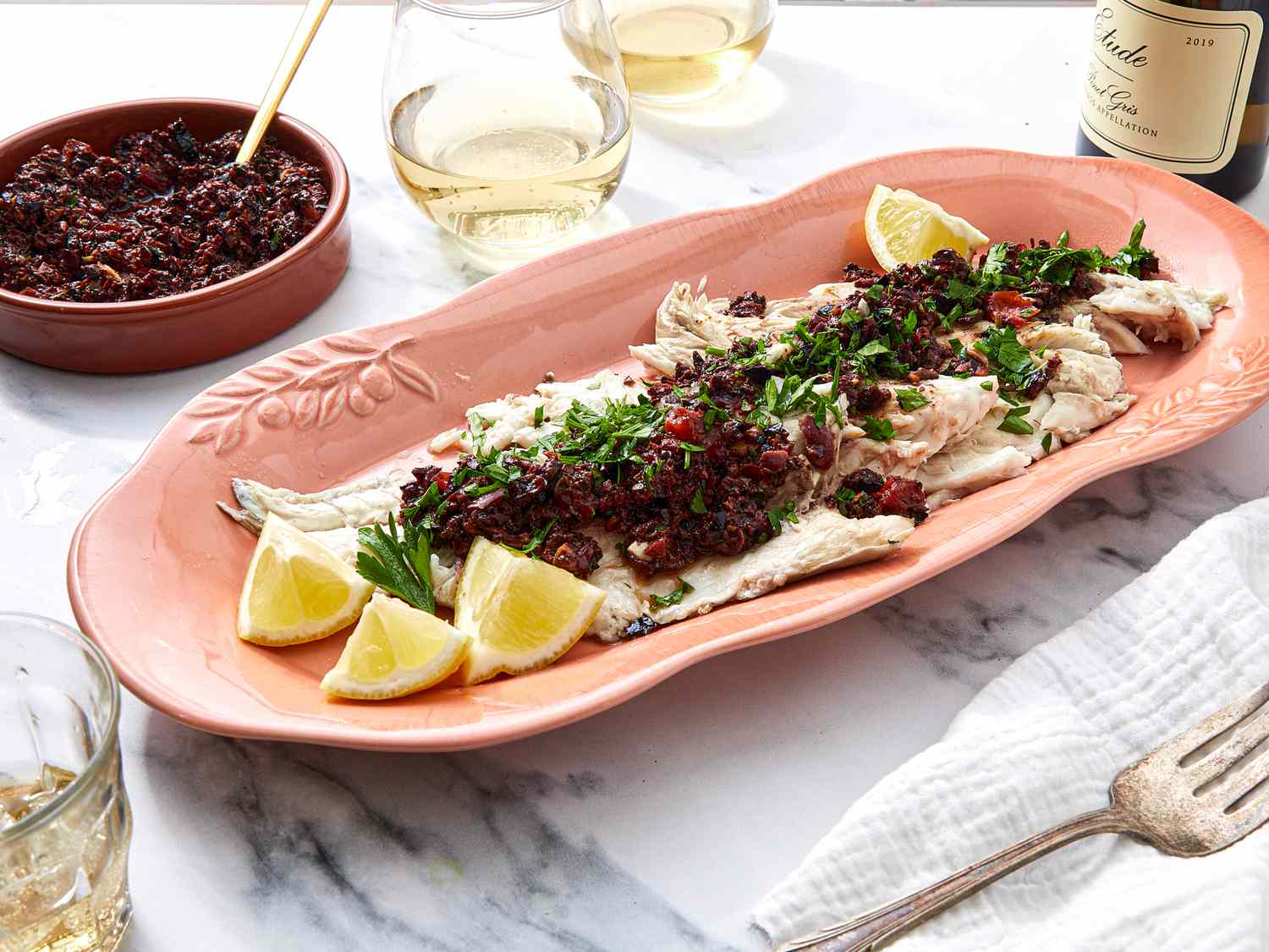 A dish of grilled fish topped with olive and tomato compote served on a table with lemon wedges and parsley garnish