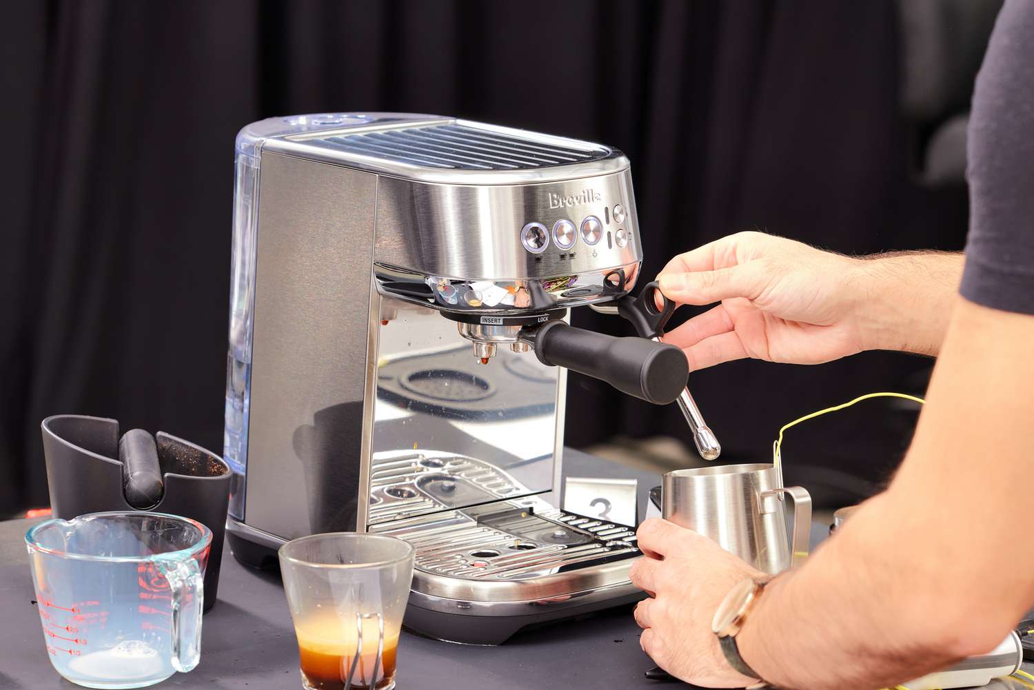 Hand using steam wand grip on Breville Bambino Plus Espresso Machine next to glass mug and measuring cup