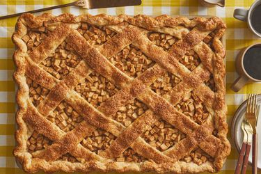 Whole Apple Slab Pie being placed on a plate. Tray is on a yellow gingham print tablecloth, with two cups of coffee, plates and gold forks 