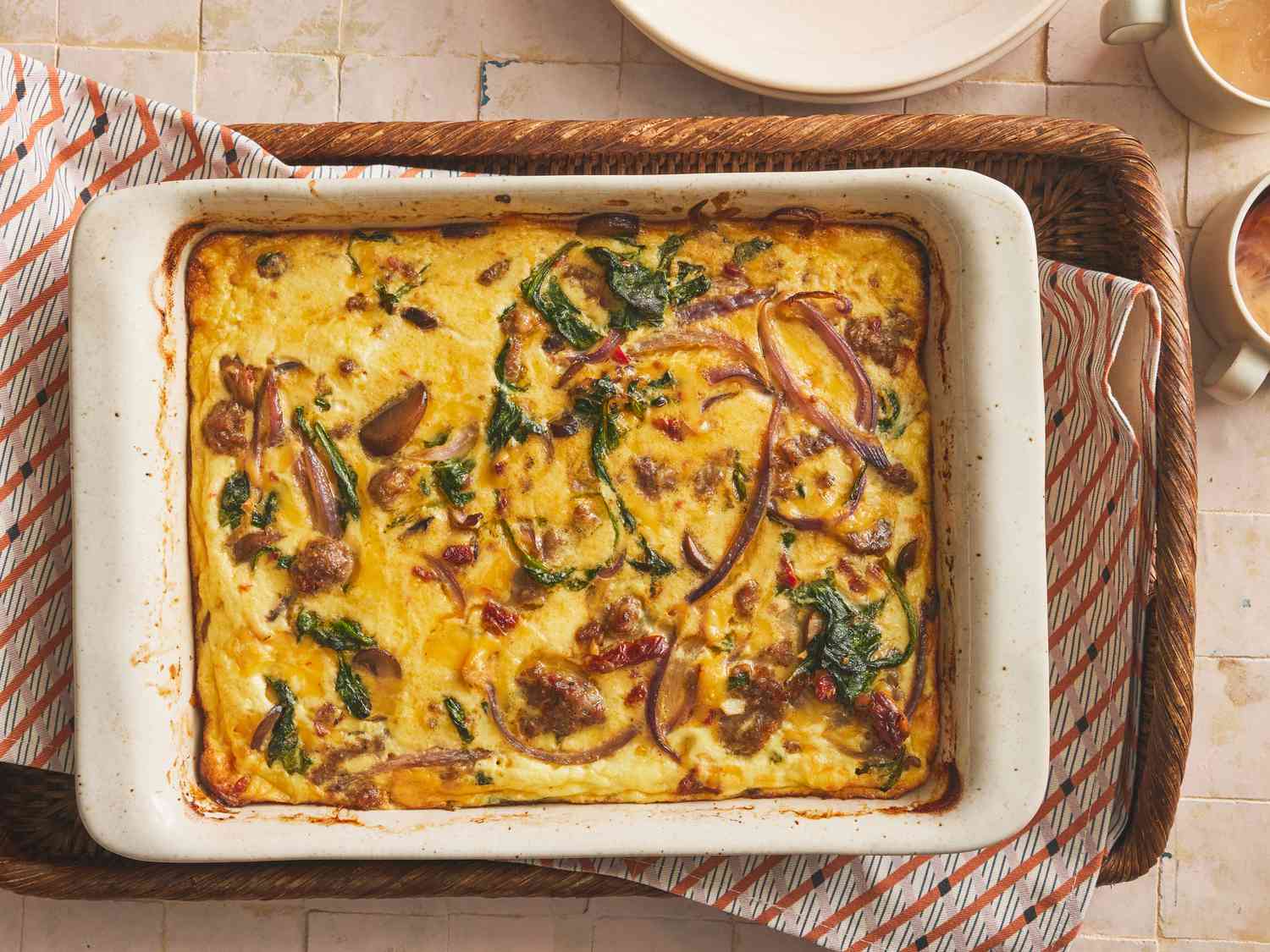 Whole breakfast casserole from above. Baking dish is in a basket lined with a striped napkin, and a rustic tiled surface. Cups of coffee and plates on the side of the frame. 