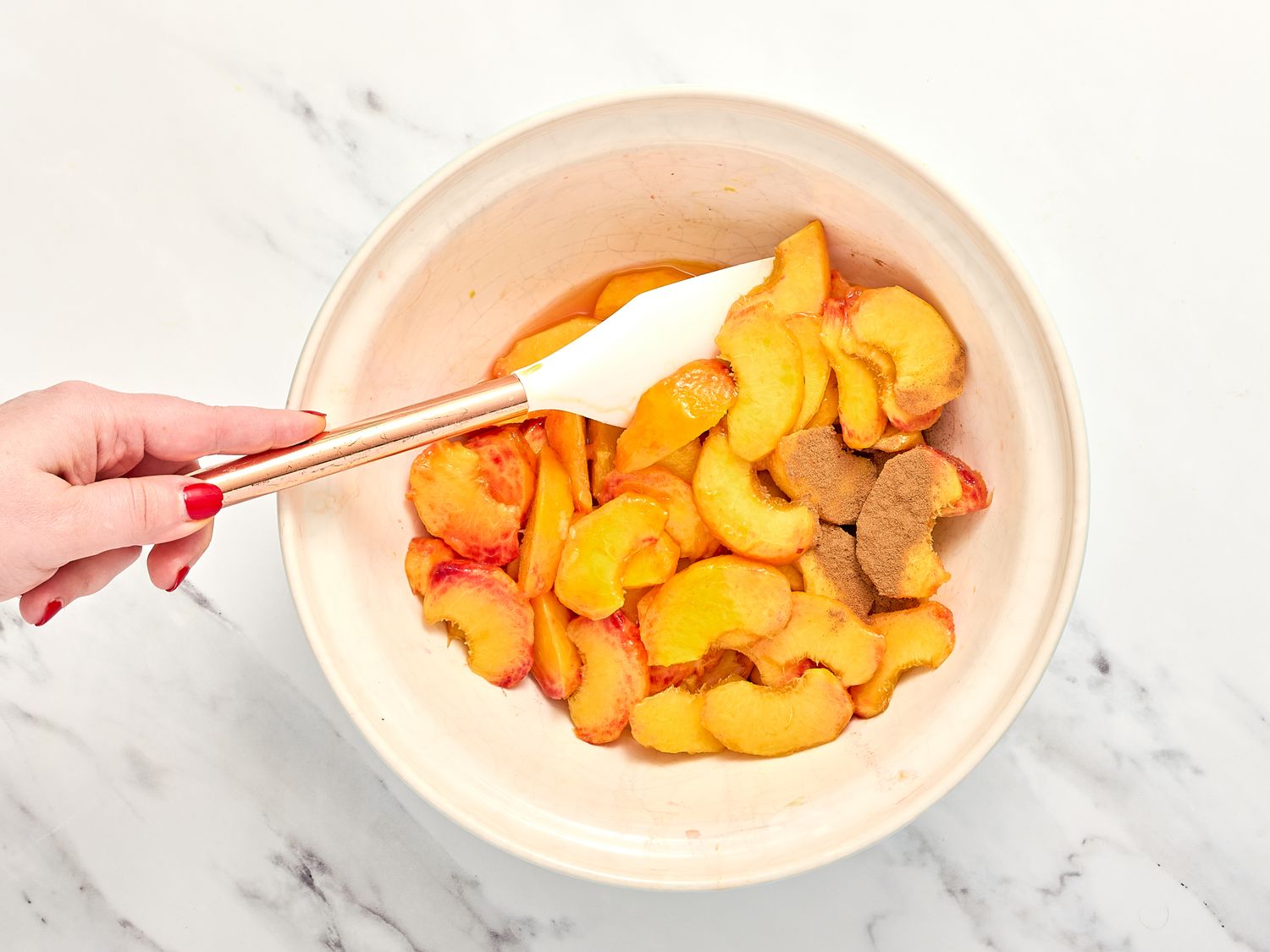 Bowl containing sliced peaches and sugar being mixed with a spatula