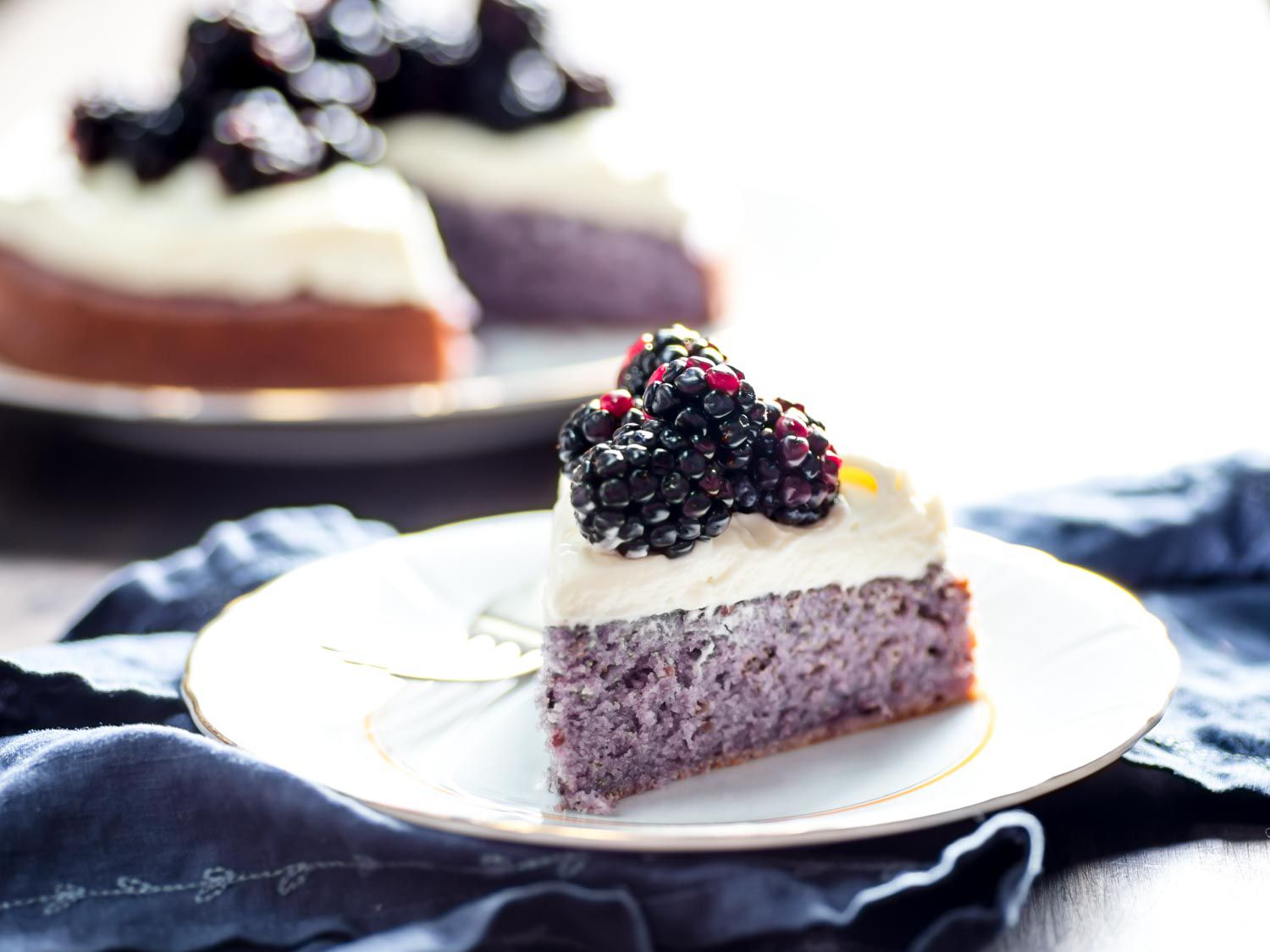 A wedge of blackberry cake on a plate, with the rest of the cake visible in the background.