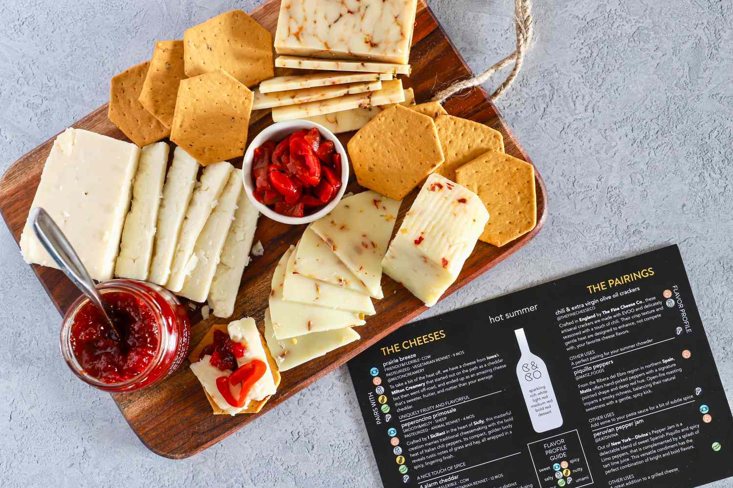 Cheese, crackers and a small bowl of peppers from Curdbox Cheese Subscription Box on a wooden board alongside a jar of red jam and a pairing menu sheet