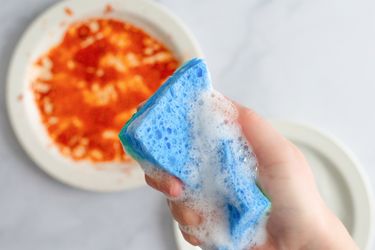 Hand holding a soapy sponge in front of a dirty plate