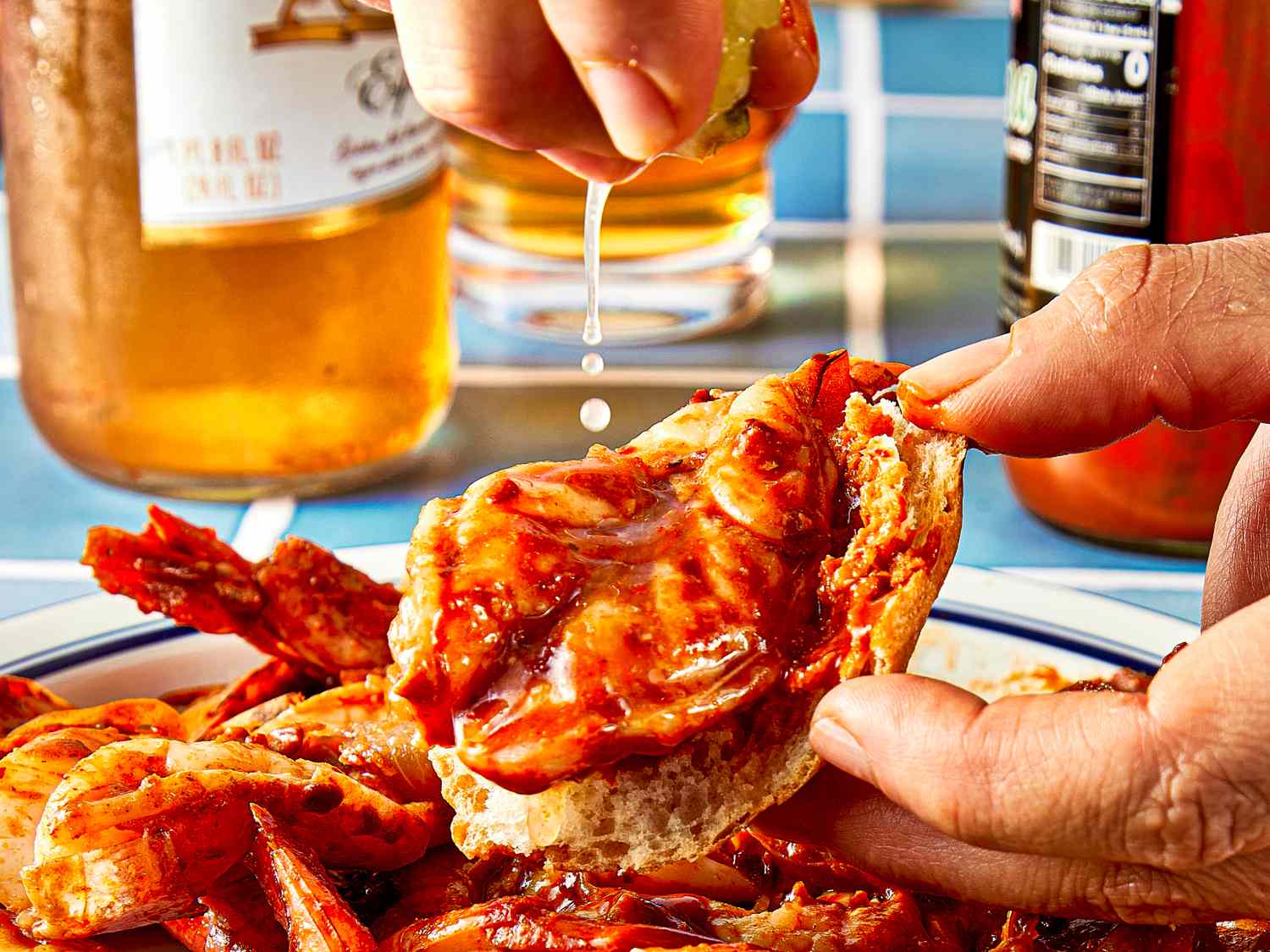 A hand squeezing lime onto a shrimp dish with sauce on a plate beer bottles in the background