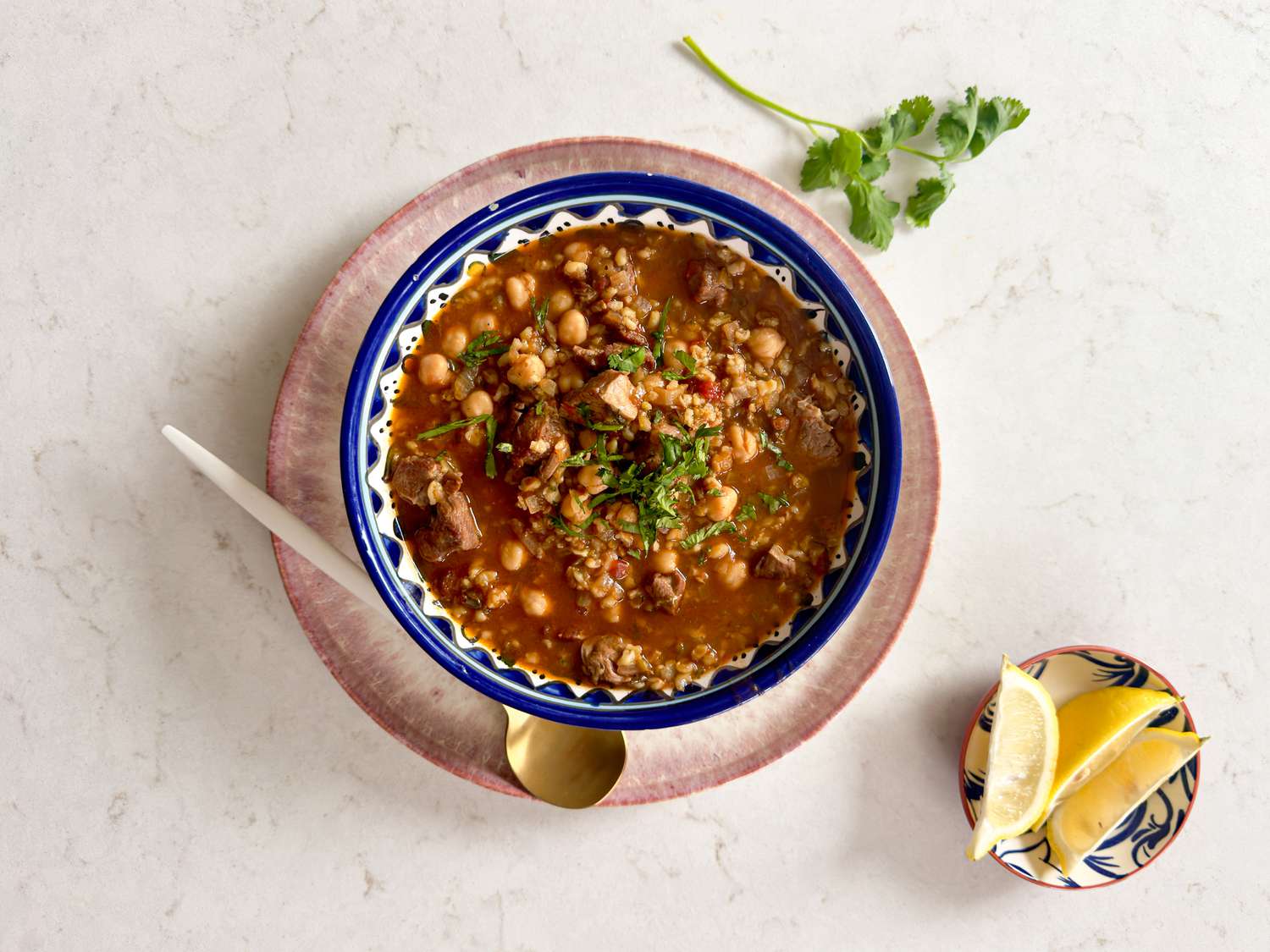 Bowl of Chobra Frik in a blue and white bowl, with small dish of lemons and cilantro on a white marble background