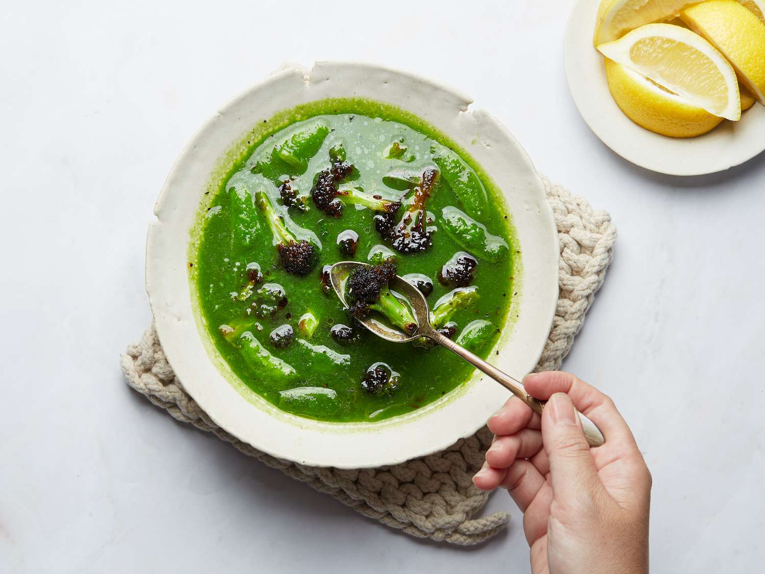 Green Broccoli, leek and snap pea soup from above on a small woven hot pad, with a person spooning a broccoli, with lemons 