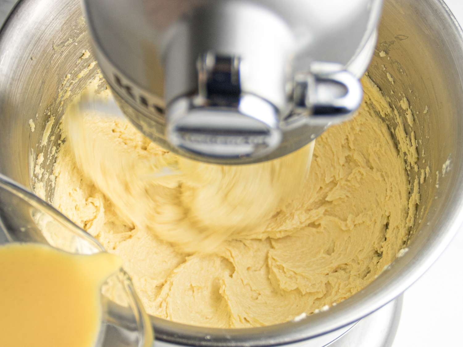 Stand mixer blending batter while liquid is poured in, process of mixing ingredients for lemon pound cake.