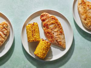 A grilled boneless chicken breast on a white ceramic plate along with two grilled pieces of corn-on-the-cob.