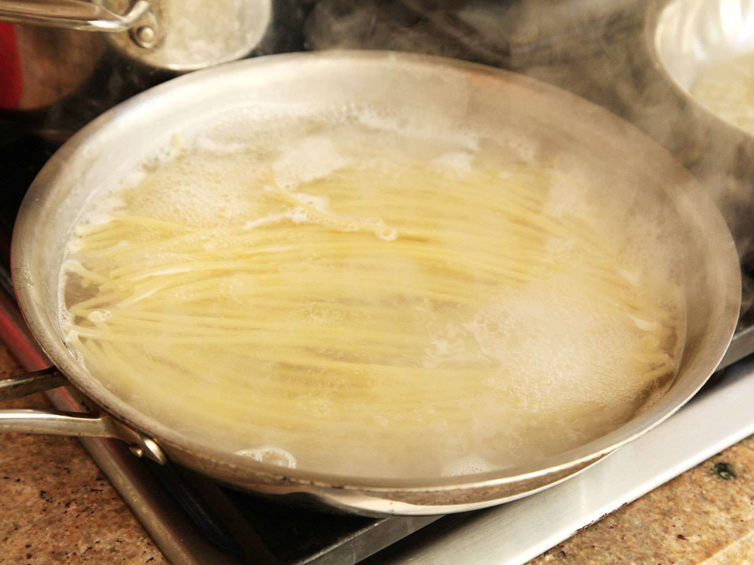 Long strands of spaghetti cooking in a small amount of water in a stainless steel skillet.