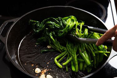 Broccoli rabe being sautéed in a cast iron skillet with a liberal amount of olive oil. Smashed garlic cloves and chile flakes are visible in the pan.