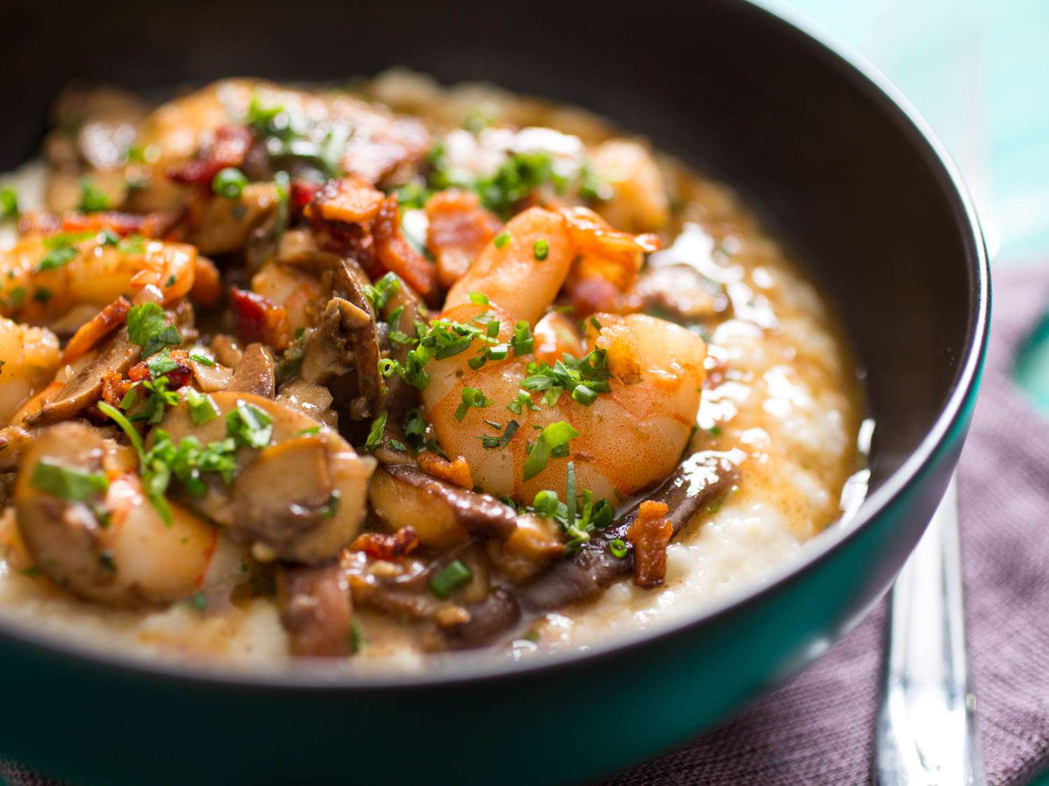 A close-up view of cheesy grits with shrimp, mushrooms, bacon, and herbs in a bowl
