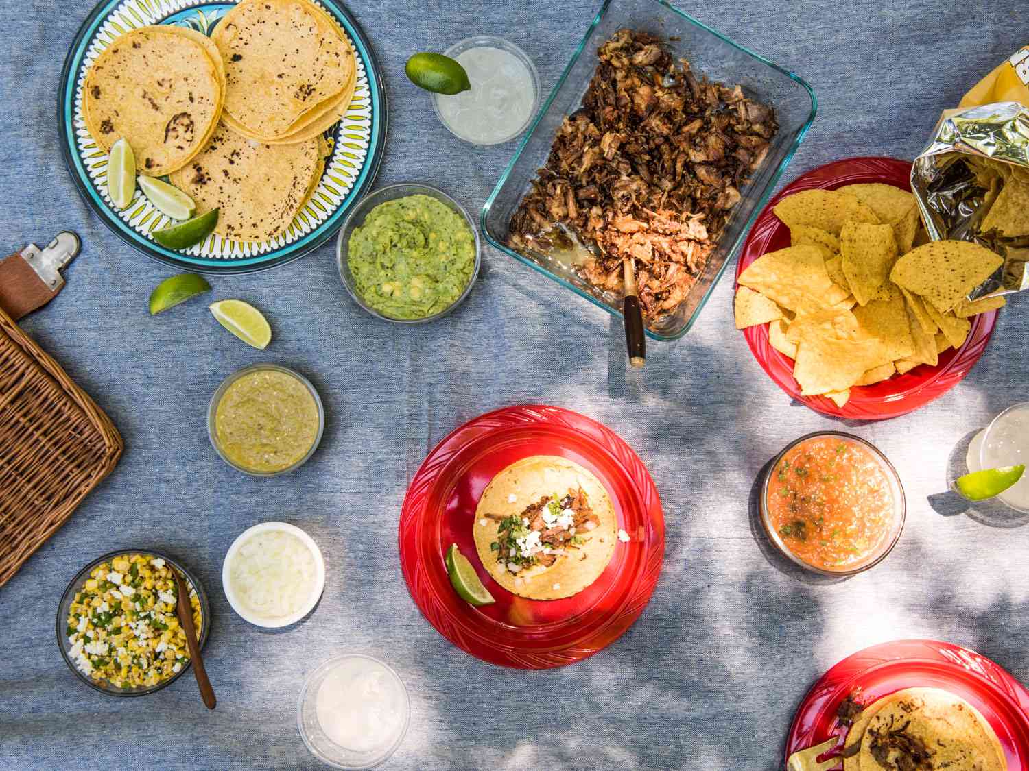 Snacks, dips, tortillas, lime wedges, salads, and dips as part of a Mexican-themed picnic spread on a picnic blanket. 