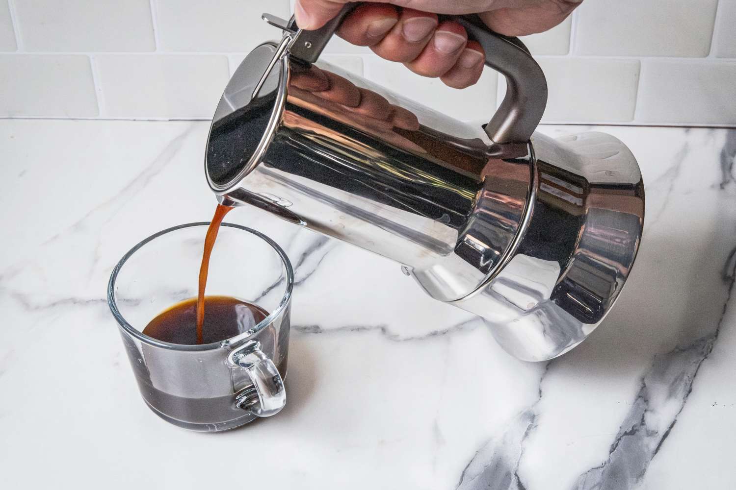 A person pouring coffee from a metal pot into a glass cup on a marble surface
