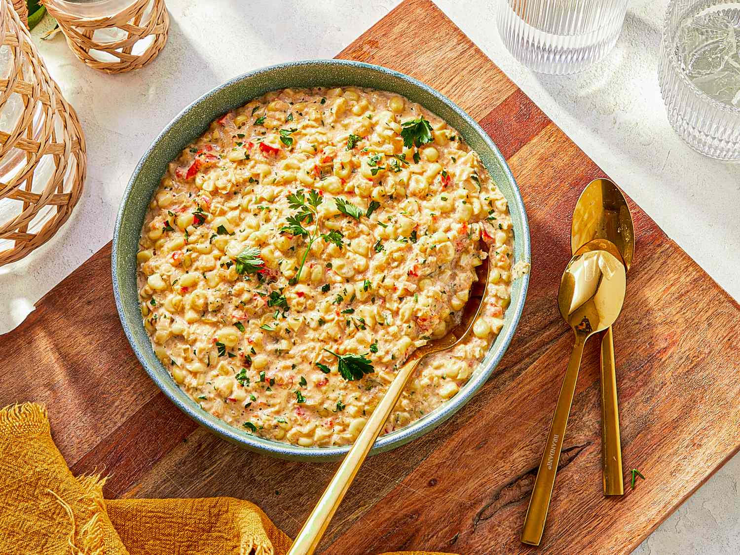 A bowl of creamy corn garnished with herbs on a wooden surface with golden utensils alongside
