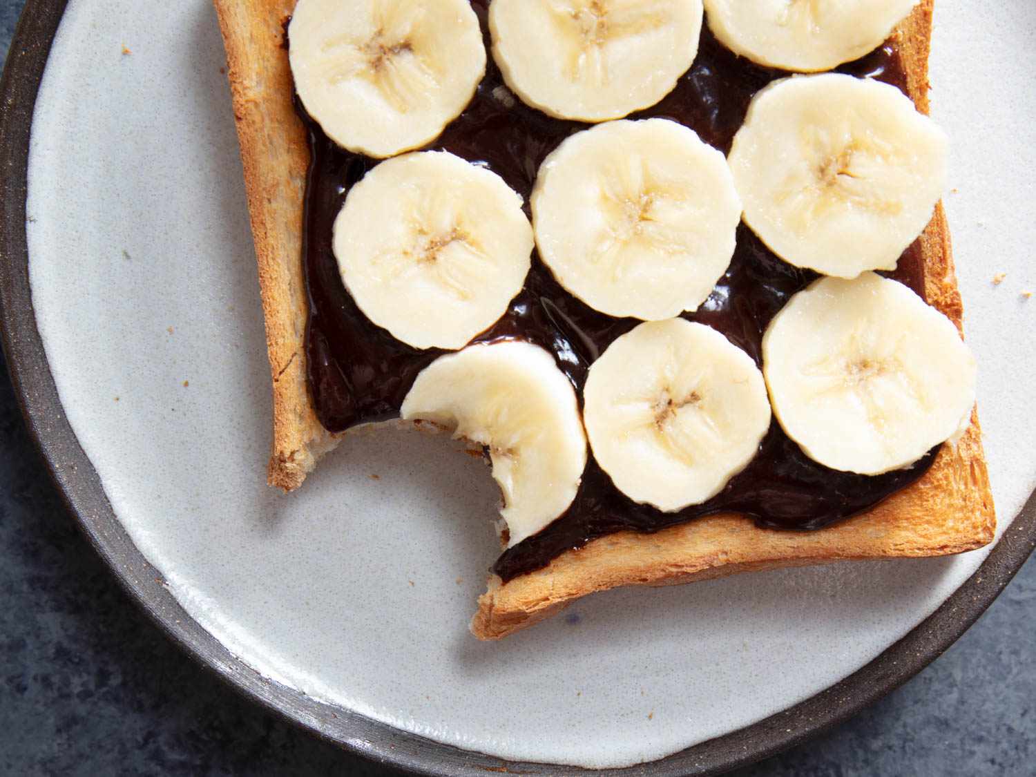 A slice of toast smeared with homemade Nutella spread, topped with sliced banana. A bite has been taken out of the toast. 