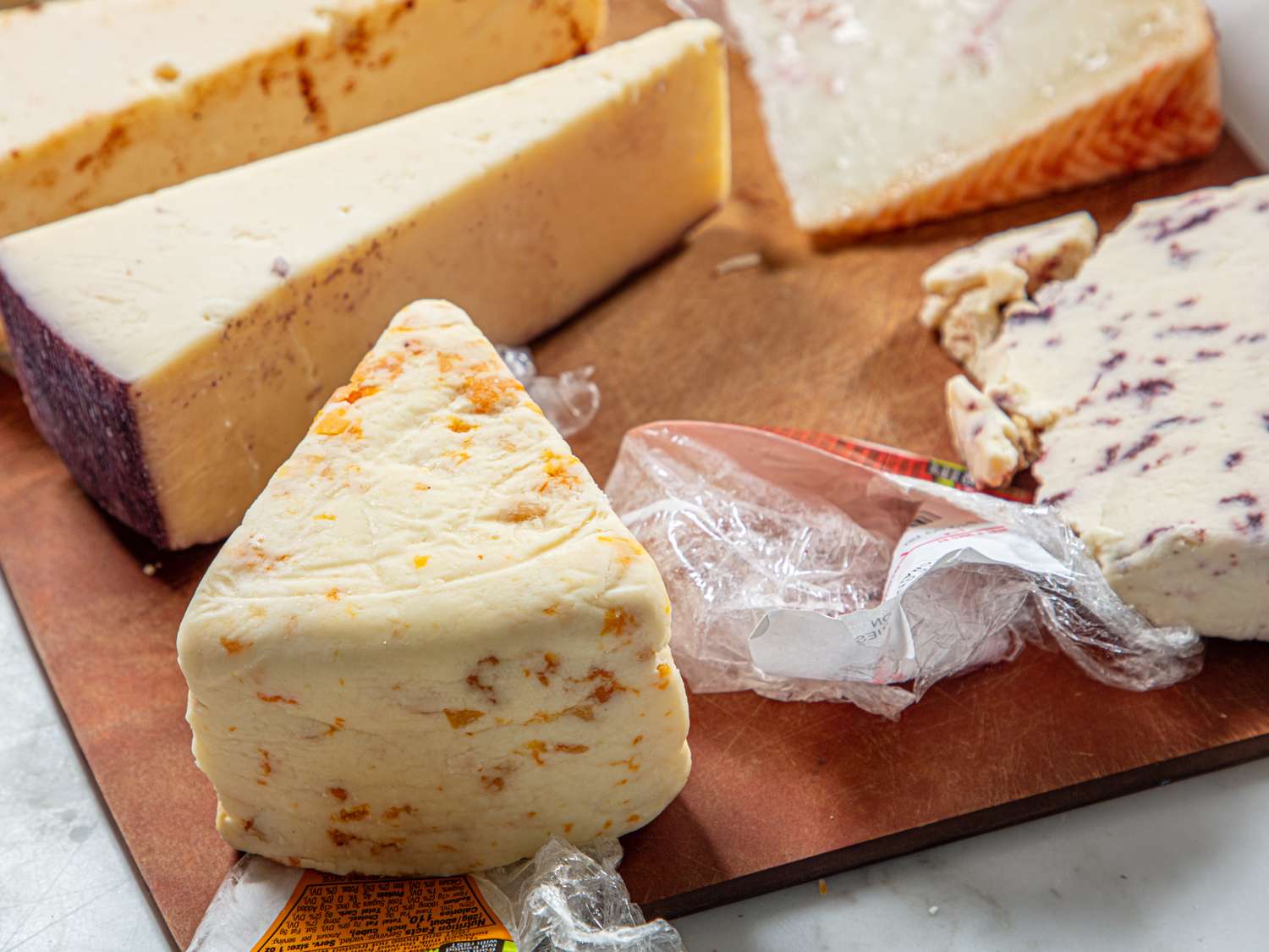 A wooden board with assorted wedges of cheese some partially wrapped in plastic