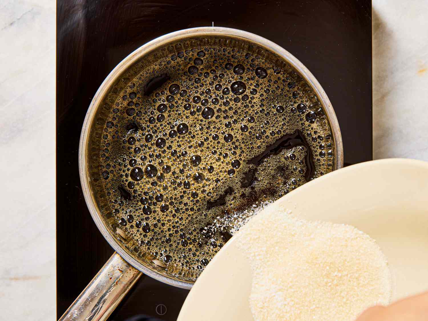 A saucepan of bubbling liquid on a hotplate with sugar or a granulated substance being poured in from a bowl