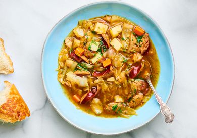 Polish potato, kielbasa, and cabbage soup in a blue bowl, with torn bread on the left periphery.