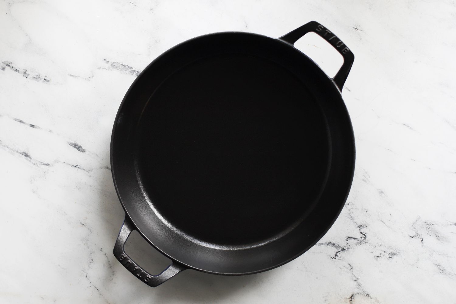 A black cast iron braiser with two handles displayed on a white marble surface