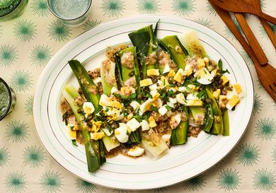 A plated dish of leeks topped with vinaigrette and chopped ingredients, displayed on a patterned surface with tableware