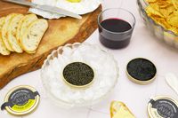 A spread on a table featuring bread slices caviar tins with a bowl of ice chips and a glass of wine