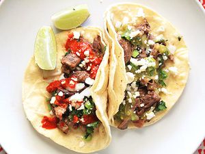 Overhead shot of two tacos de lengua on a plate. One is garnished with a red salsa and the other sports green salsa. Both are sprinkled with queso fresco, scallions, and cilantro. Two lime wedges flank the tacos.