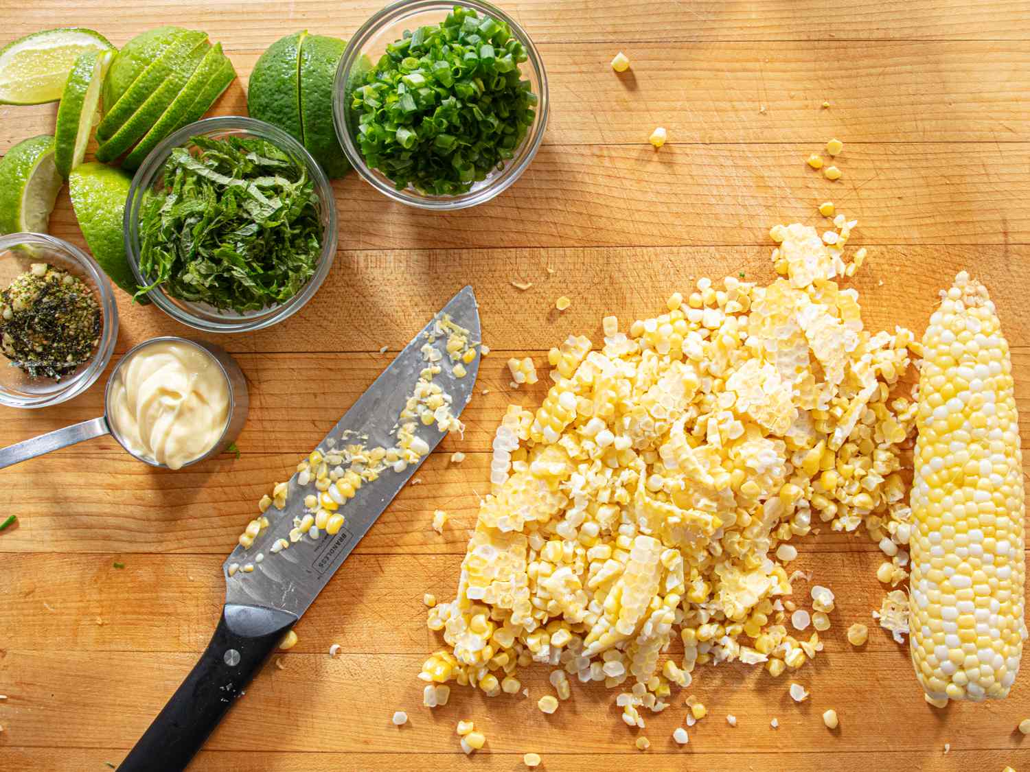 Ingredients for a corn salad on a wooden surface including corn herbs lime slices and a knife