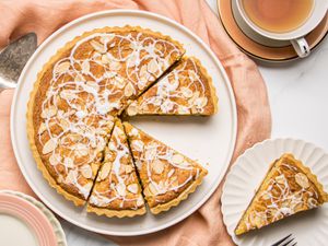 A bakewell tart served on a plate, sliced with one piece on a separate dish next to a cup of tea