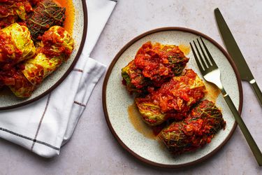On the right of the images is three stuffed cabbage rolls topped with tomato sauce on a ceramic plate with metal flatware. On the left hand side of the image is a serving platter holding more cabbage rolls which is set on a folded cloth.